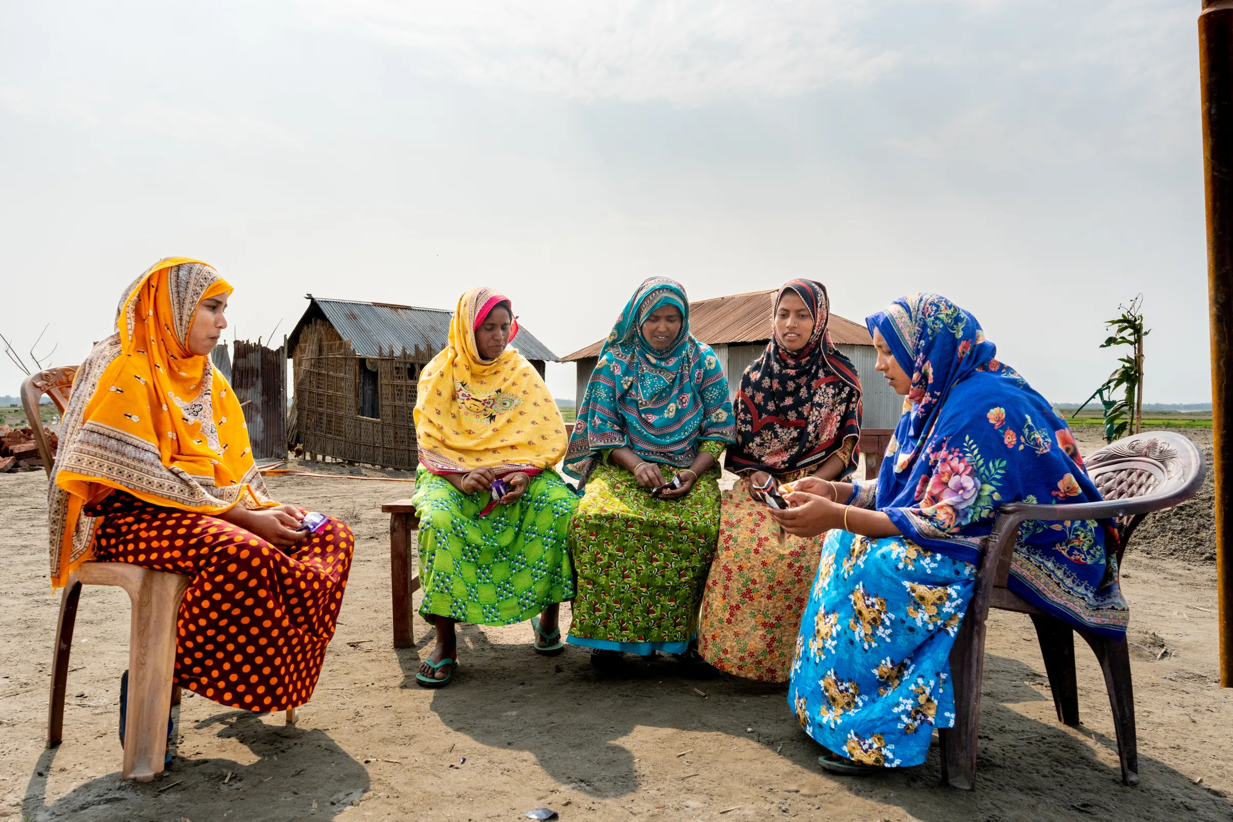 A group of 5 Bangladeshi women, colourfully dressed sitting on chairs outside 