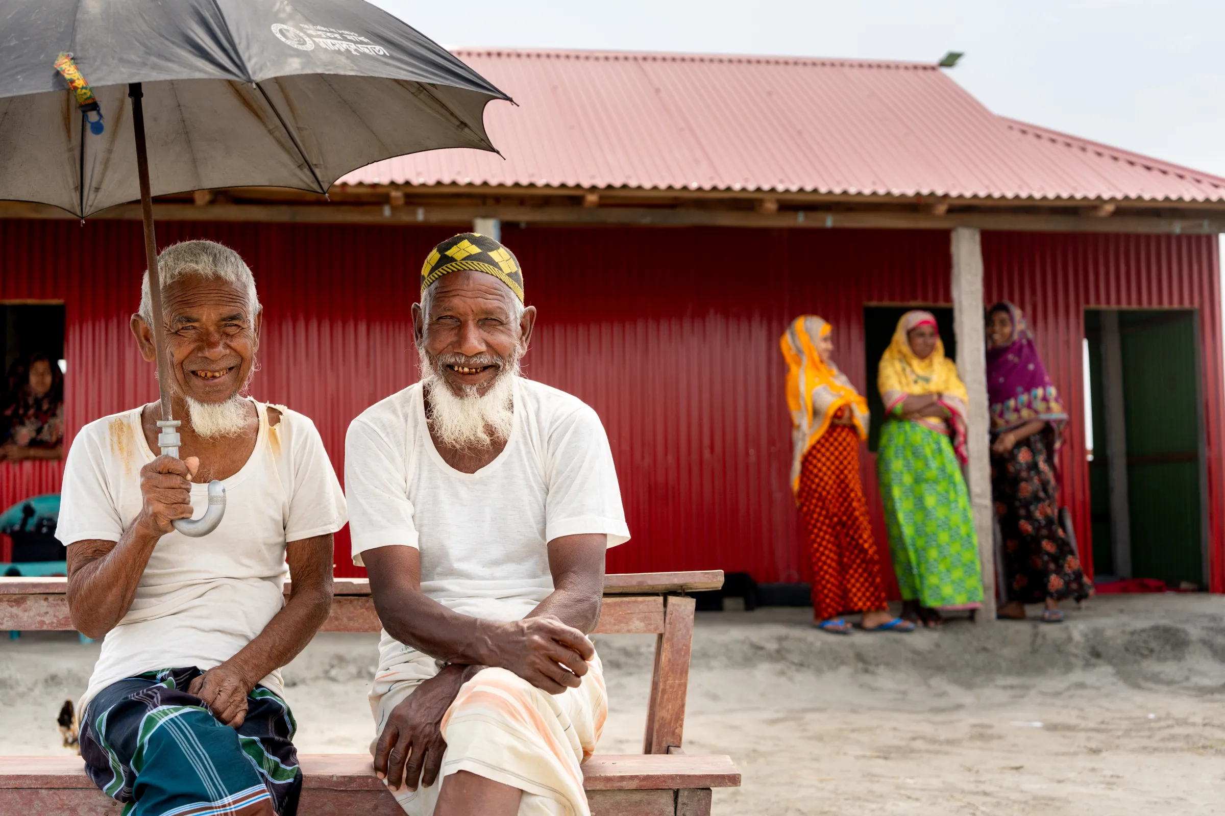 Two Bangladeshi men sitting outside under an umbrella, three women standing in the background