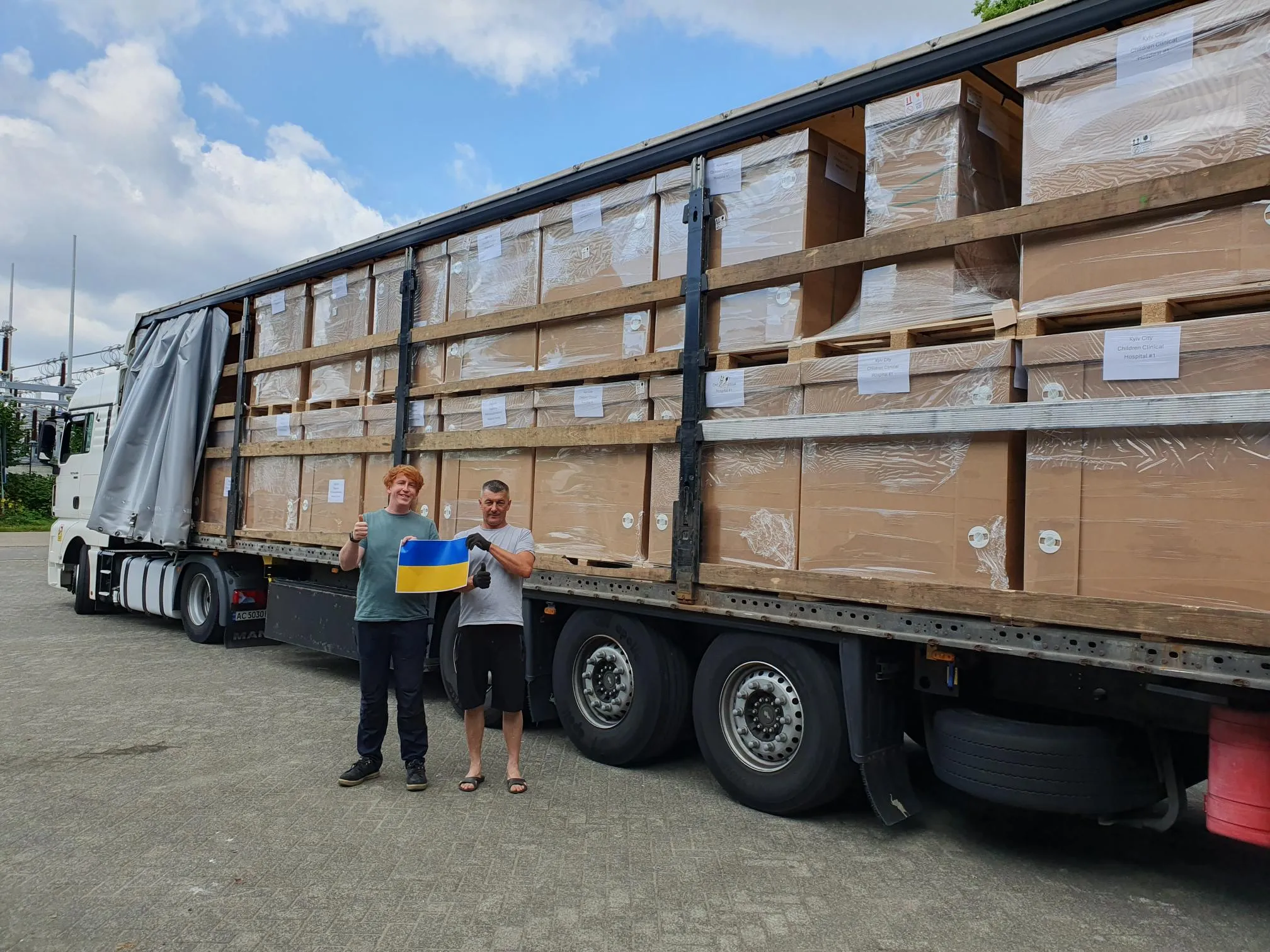 A truck loaded with boxes containing solar panels. In front of the truck, two proud truck drivers holding the Ukrainian flag.